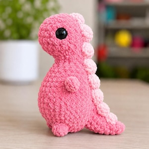 Könnte beinhalten: Ein kleines, handgefertigtes Häkel-Dinosaurier-Stofftier in leuchtendem Pink. Die Figur hat einen runden Körper, einen großen Kopf mit einem sichtbaren schwarzen Kunststoffauge und eine Reihe hellrosa, abgerundeter Stacheln am Rücken.