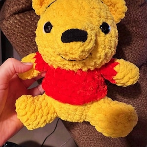 May include: A yellow crocheted bear plush toy with a textured, fuzzy appearance. The bear features black plastic eyes, a black stitched nose, and a small stitched mouth. It is wearing a bright red crocheted shirt that covers its torso.