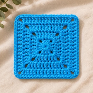 May include: A square crochet coaster in a vibrant bright blue color. The piece features a classic granny square pattern with a central cluster of stitches, radiating outward to form a geometric shape with openwork spaces between the yarn rows.