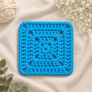May include: A square crochet coaster in a vibrant bright blue color. The piece features a classic granny square pattern with visible stitches and openwork gaps arranged in a symmetrical design, showcasing a consistent texture throughout the item.