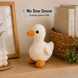 May include: A handmade, white crochet goose with an orange beak and feet. The goose is sitting on a wooden shelf next to a framed picture and a decorative candle. The text "No Sew Goose Crochet pattern PDF" is visible.