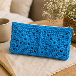 May include: A rectangular crochet pouch in a vibrant blue color, featuring two prominent granny square motifs on the front. The pouch is secured with a gold-toned metal zipper closure that includes a small, round, metallic charm pull attached.