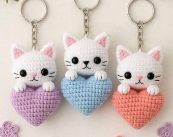 May include: Three crochet keychains featuring white cats with black eyes and pink inner ears. Each cat holds a heart-shaped base in lavender, light blue, or peach. The keychains include a silver-toned metal ring and chain attached to the top of each cat.