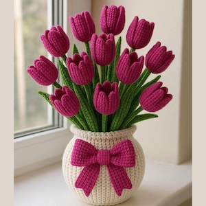 Könnte beinhalten: Ein handgefertigter Häkelstrauß aus magentafarbenen Tulpen in einer cremefarbenen Vase. Die Vase hat eine magentafarbene Schleife. Die Tulpen haben grüne Stiele und Blätter. Das Arrangement wird auf einer Fensterbank ausgestellt.