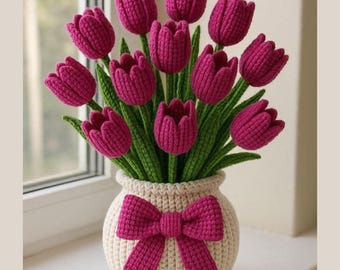 Könnte beinhalten: Ein handgefertigter Häkelstrauß aus magentafarbenen Tulpen in einer cremefarbenen Vase. Die Vase hat eine magentafarbene Schleife. Die Tulpen haben grüne Stiele und Blätter. Das Arrangement wird auf einer Fensterbank ausgestellt.