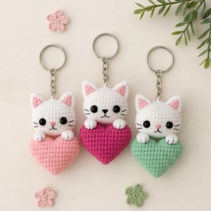 May include: Three crochet cat keychains, each holding a heart. The cats are white with pink accents, black eyes, and whiskers. The hearts are pink, fuchsia, and green. Each keychain has a silver ring.