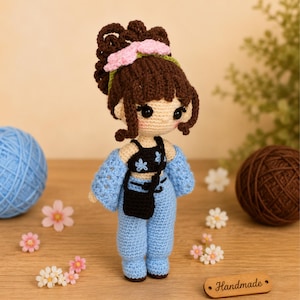 Puede incluir: Una muñeca de ganchillo con cabello castaño peinado en un moño alto con un accesorio de flor rosa. La muñeca viste un top corto negro con detalles de flores azules, un bolso cruzado negro y pantalones azul claro con una prenda azul a juego.