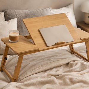 May include: A light brown bamboo laptop desk with adjustable legs and a tilting surface. A silver laptop rests on the tilted surface. A glass of coffee sits on the side. The desk is on a bed with white and grey pillows.