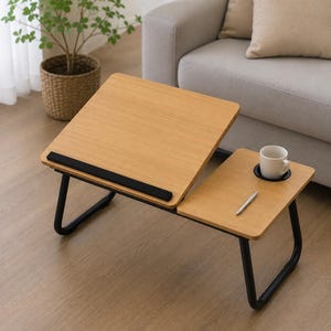May include: A portable laptop desk with a light wood grain finish and black metal legs. The desk features an adjustable tilting surface with a black ledge, a fixed side section with a circular cup holder, a white ceramic mug, and a silver pen.
