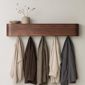 Peut inclure: Étagère murale en bois foncé avec cinq crochets, présentant cinq vestes de couleurs neutres. Un petit vase avec des fleurs séchées et un bol sont posés sur l'étagère. Le tout est fixé sur un mur neutre.