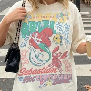 Może przedstawiać: Kremowa koszulka z kolorową grafiką Arielki, Florka i Sebastiana z Małej Syrenki, z napisem "ARIEL OCEAN TOUR 1991" i "Sebastian with the Hot Crustacean Band". Koszulka jest noszona z beżową czapką bejsbolową.