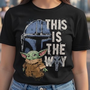 May include: Black t-shirt with a graphic of a Mandalorian helmet, Baby Yoda, and the text "THIS IS THE WAY". The design includes a helmet and a small figure in a brown coat. A castle is visible in the background.
