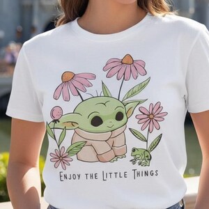 May include: White t-shirt featuring a green cartoon character with large eyes, surrounded by pink flowers and a small green frog. The text "Enjoy the Little Things" is printed below the graphic. The background includes a castle.