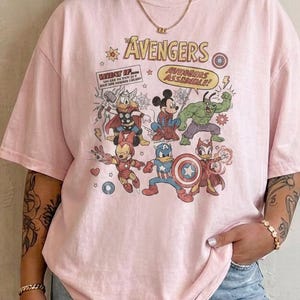May include: A light pink short-sleeve t-shirt featuring a graphic print of cartoon characters dressed as The Avengers. The text reads The Avengers and Avengers Assemble. The design includes illustrations of characters resembling Thor, Hulk, Iron Man, and Captain America.