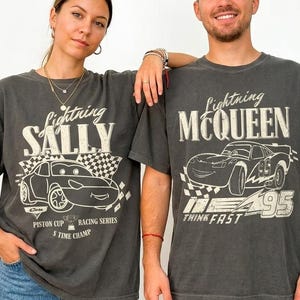 May include: Two charcoal gray t-shirts with Lightning McQueen and Sally designs. The Lightning McQueen shirt has the text "Think Fast" and "95". The Sally shirt has the text "Piston Cup Racing Series" and "5 Time Champ".