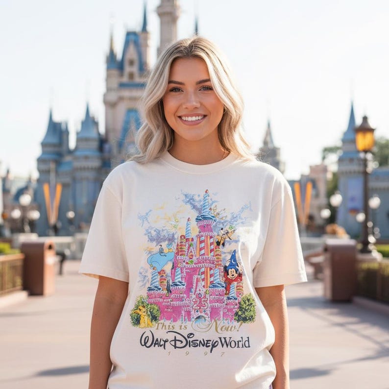 May include: A cream-colored short-sleeve t-shirt featuring a colorful graphic of a pink castle with Disney characters, the text "This is Now!" and "Walt Disney World 1997" printed on the front, showcasing a 25th-anniversary design theme.