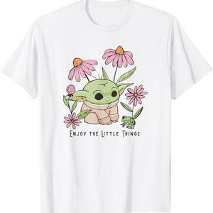 May include: White t-shirt featuring a cartoon character with large eyes, surrounded by pink flowers and green leaves. The text "ENJOY THE LITTLE THINGS" is printed below the character.