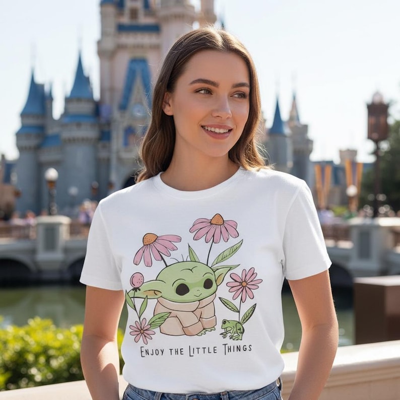 May include: White t-shirt featuring a green cartoon character with large eyes, surrounded by pink flowers and a small green frog. The text "Enjoy the Little Things" is printed below the graphic. The background includes a castle.