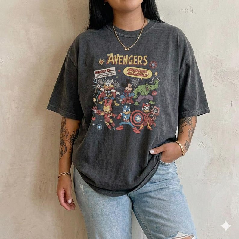 May include: A charcoal grey short-sleeve t-shirt featuring a graphic print of cartoon characters dressed as Avengers. The text reads The Avengers and Avengers Assemble. The design includes illustrations of characters resembling Thor, Mickey Mouse, Hulk, Iron Man, Captain America, and Donald Duck.
