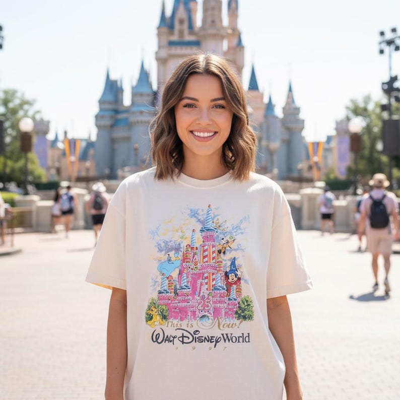 May include: A cream-colored short-sleeve t-shirt featuring a colorful graphic of a castle with the text "25", "This is the Magic", and "Walt Disney World 1997" printed on the front. The graphic includes illustrations of various cartoon characters.