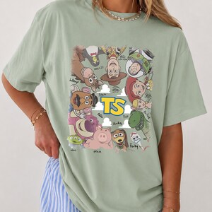 May include: A sage green short-sleeve t-shirt featuring a graphic print of various animated characters arranged in a circle around the yellow text TS. The characters include a cowboy, space ranger, cowgirl, dinosaur, pig, and other figures.