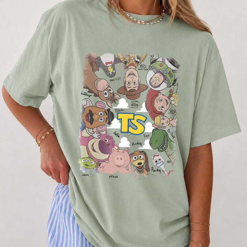 May include: A sage green short-sleeve t-shirt featuring a graphic print of various animated characters arranged in a circle around the yellow text TS. The characters include a cowboy, space ranger, cowgirl, dinosaur, pig, and other figures.