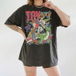 May include: Dark gray t-shirt featuring the Toy Story logo and characters Woody, Buzz Lightyear, and Rex. The shirt has a vintage look with the text "Since 1995". The design includes red, blue, green, and yellow colors.