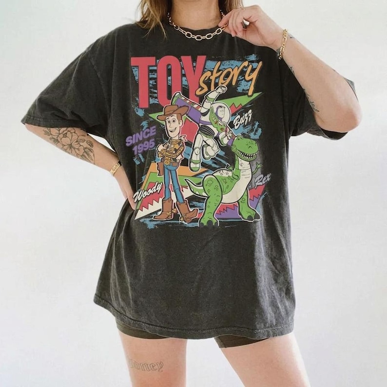 May include: Dark gray t-shirt featuring the Toy Story logo and characters Woody, Buzz Lightyear, and Rex. The shirt has a vintage look with the text "Since 1995". The design includes red, blue, green, and yellow colors.