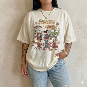May include: An off-white short-sleeve t-shirt featuring a graphic print of cartoon characters dressed as The Avengers. The text reads The Avengers and Avengers Assemble. The design includes various comic book style illustrations of characters.