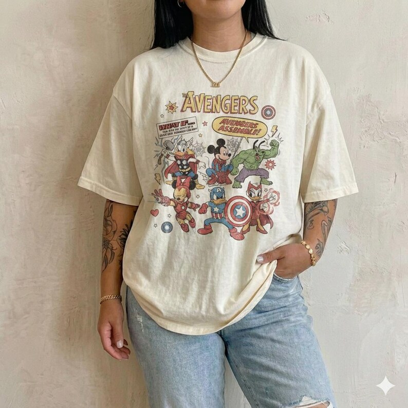 May include: An off-white short-sleeve t-shirt featuring a graphic print of cartoon characters dressed as The Avengers. The text reads The Avengers and Avengers Assemble. The design includes various comic book style illustrations of characters.