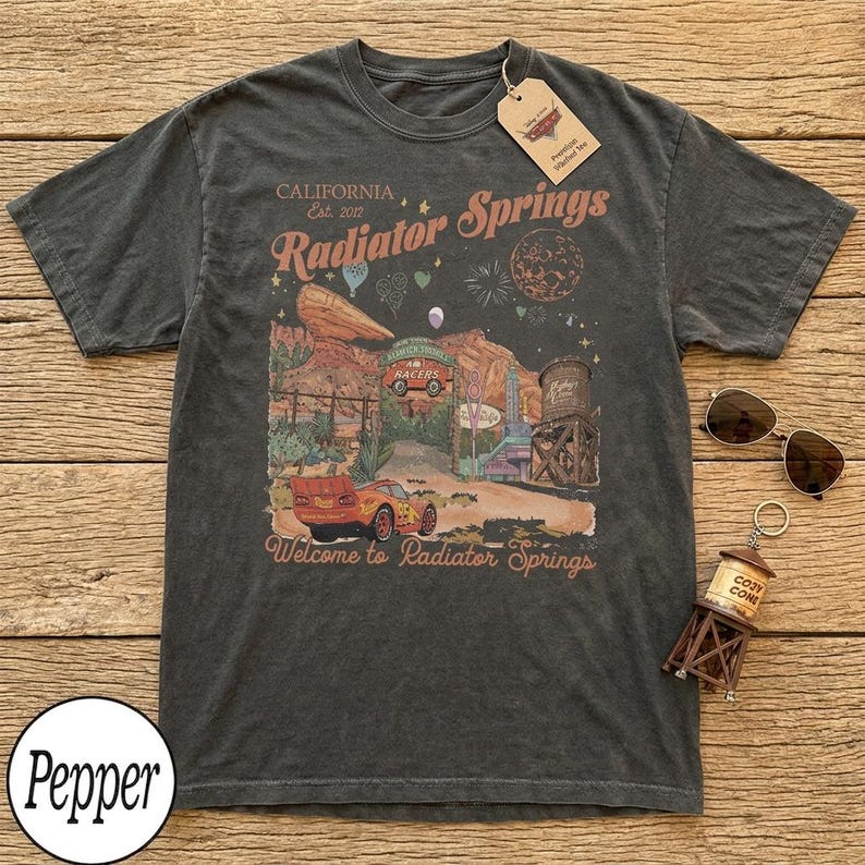 May include: Dark gray t-shirt featuring a colorful graphic of Radiator Springs, with text that reads "California Est. 2012" and "Welcome to Radiator Springs." The design includes a car, desert landscape, and water tower.