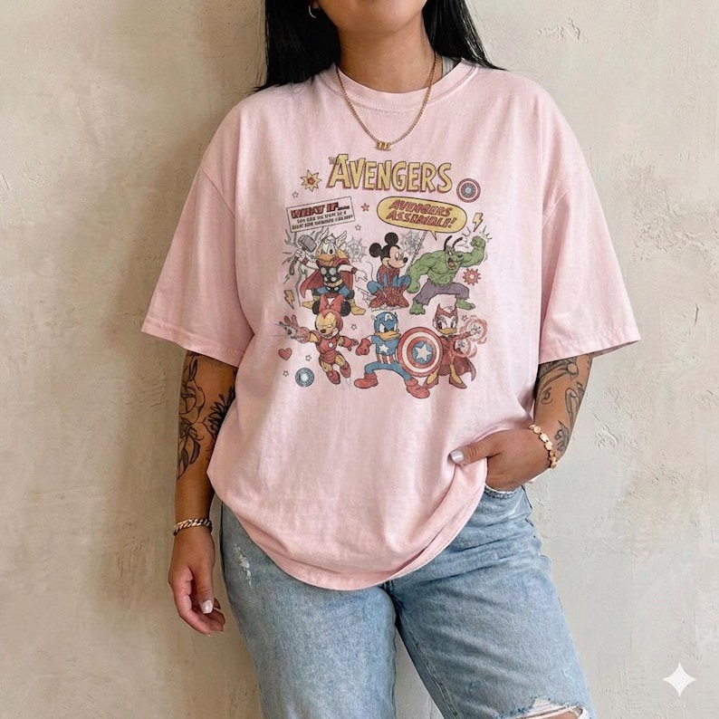 May include: A light pink short-sleeve t-shirt featuring a graphic print of cartoon characters dressed as The Avengers. The text reads The Avengers and Avengers Assemble. The design includes illustrations of characters resembling Thor, Hulk, Iron Man, and Captain America.