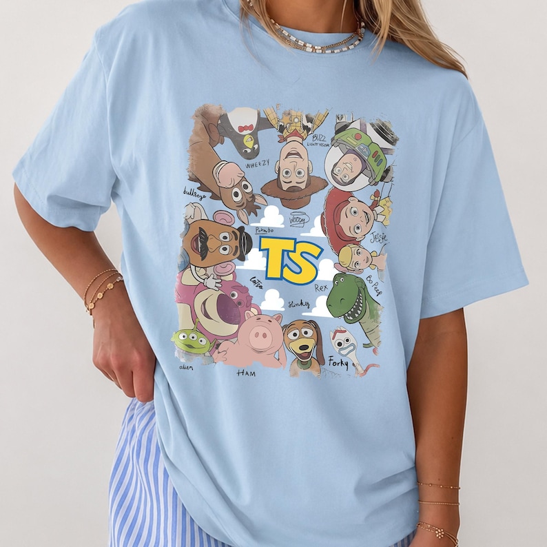 May include: A light blue t-shirt featuring a graphic print of various animated characters arranged in a circle around the yellow text TS. The characters include a cowboy, space ranger, cowgirl, dinosaur, pig, and other figures with their names written below.