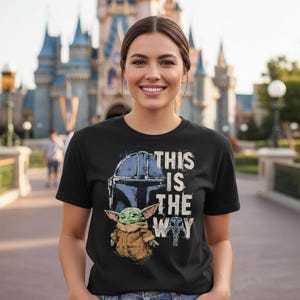 May include: Black t-shirt with a graphic of a Mandalorian helmet, Grogu, and the phrase "This is the Way" in white. The design includes a blue helmet and Grogu in a brown coat. The background shows a castle.