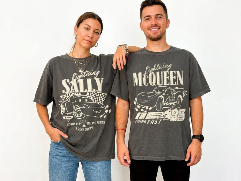 May include: Two charcoal gray t-shirts with Lightning McQueen and Sally designs. The Lightning McQueen shirt has the text "Think Fast" and "95". The Sally shirt has the text "Piston Cup Racing Series" and "5 Time Champ".