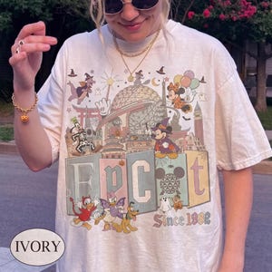 Puede incluir: Una camiseta de manga corta color marfil con un estampado gráfico colorido de personajes de Disney con disfraces de Halloween, incluyendo a Mickey y Minnie Mouse, Goofy y Donald Duck, con el texto EPCOT Since 1982 y Spaceship Earth.