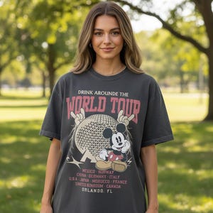 Op de afbeelding: Donkergrijs t-shirt met de tekst "DRINK AROUND THE WORLD TOUR" in roze en wit. Het shirt heeft een afbeelding van Mickey Mouse en een wereldbol. Verschillende landen en Orlando, FL staan op het shirt.