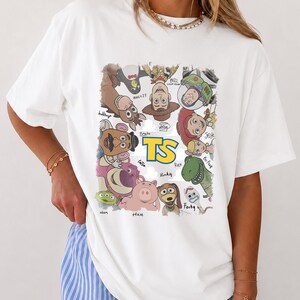 May include: A white t-shirt featuring a colorful graphic print of various animated characters arranged in a circle around the yellow text TS. The characters include a cowboy, space ranger, cowgirl, dinosaur, pig, and others with their names written below.