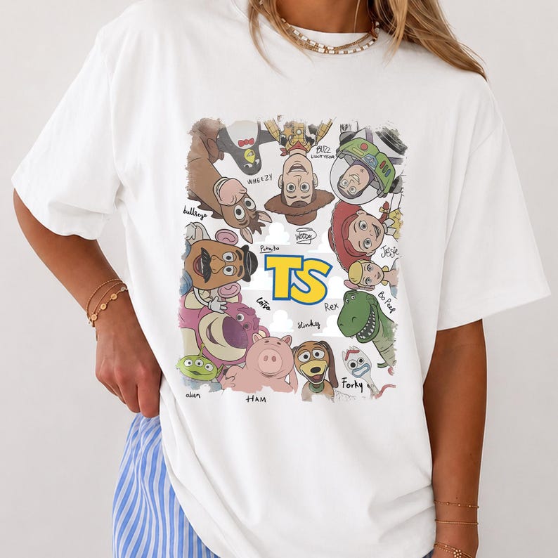 May include: A white t-shirt featuring a colorful graphic print of various animated characters arranged in a circle around the yellow text TS. The characters include a cowboy, space ranger, cowgirl, dinosaur, pig, and others with their names written below.