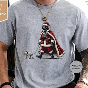 Pode incluir: Uma t-shirt de manga curta em cinzento mesclado atlético com um gráfico de uma personagem vestida de Pai Natal a passear um pequeno andador robótico com uma trela. A personagem usa um fato, uma capa e um gorro de Pai Natal vermelho e branco.