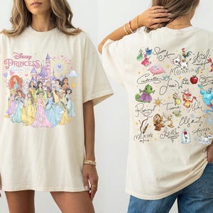 May include: Cream-colored t-shirt featuring a Disney Princess design on the front and character signatures on the back. The front graphic includes a castle, fireworks, and various princesses. The back displays character illustrations and autographs.