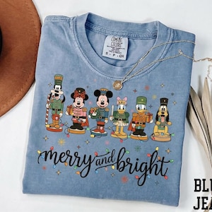 May include: A blue jean colored t-shirt featuring a graphic of six cartoon characters dressed as nutcrackers, including Goofy, Minnie Mouse, Mickey Mouse, Daisy Duck, Donald Duck, and Pluto. The text merry and bright is printed below.