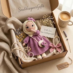 Può includere: Una bambola all'uncinetto fatta a mano in una scatola regalo. La bambola indossa un vestito viola, un cappuccio bianco e una piccola borsa. La scatola contiene una tazza di caffè, fiori e una carta con "With Love from SouldesignerGifts".