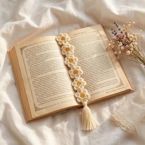 Puede incluir: Marcapáginas de ganchillo hecho a mano con flores de margarita con centros amarillos. El marcapáginas crema tiene una borla y está en un libro abierto con el texto "VINTAGE READS" y "THE COZY DAISY HANDMADE".