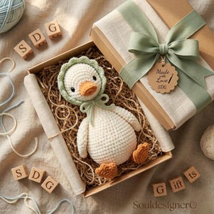 Può includere: Un giocattolo a forma di anatra all'uncinetto fatto a mano con becco e zampe arancioni, con un cappuccio e un fiocco verdi, presentato in una confezione regalo con un nastro verde. La scatola contiene il testo "Made with Love SDG".