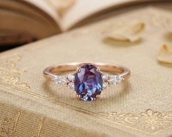May include: A rose gold ring featuring a central oval-cut color-changing alexandrite gemstone. The stone is flanked by two small round diamonds and two marquise-cut diamonds on each side, set in a delicate band with a polished metallic finish.
