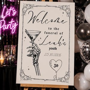 May include: A rectangular white sign on a wooden easel featuring black ink illustrations and text. The sign reads: Welcome to the funeral of Leah's youth 25.10.2026. It depicts a skeletal hand holding a martini glass and a heart-shaped balloon with 30.