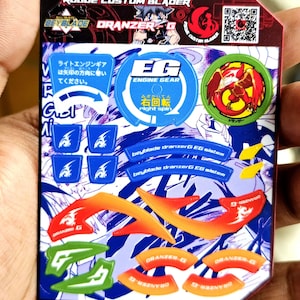 May include: A rectangular sheet of stickers for a Beyblade Dranzer G toy, featuring various blue, orange, and green decals. The top includes the text Rogue Custom Blader, Dranzer G, and Engine Gear, alongside a QR code and a circular phoenix emblem.