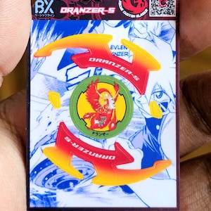 May include: A rectangular Beyblade X card featuring the text Rogue Custom Blader, Beyblade, and Dranzer-S. The card displays a central green circular emblem with a red phoenix illustration, surrounded by yellow and red arrow-shaped graphics.