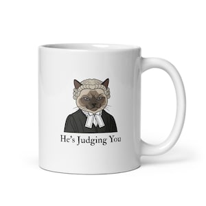 Pode incluir: Caneca de cerâmica branca com um gato de desenho animado vestido de juiz, usando peruca e toga. O gato tem uma expressão séria. O texto "He's Judging You" está impresso abaixo da imagem.